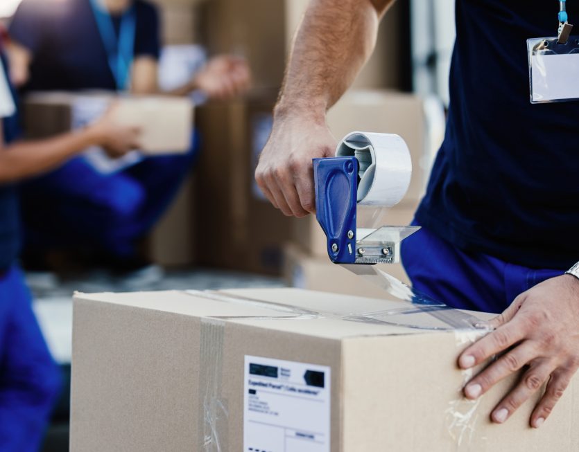 Close-up of worker using tape dispenser while packing cardboard ERP Distribution Athéna Conseil pour optimiser la supply chain et la logistique
